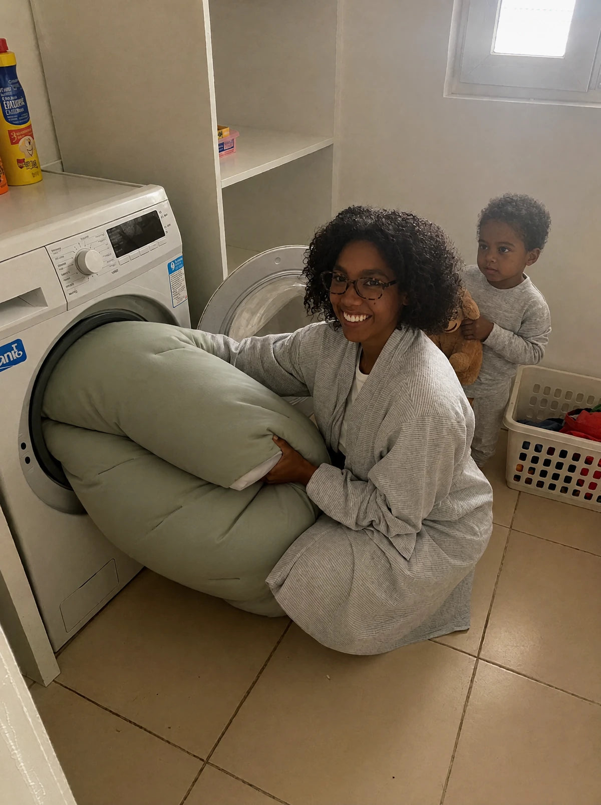 Une maman met sa couette Whoosh vert sauge entière dans le lave-linge, son fils à côté