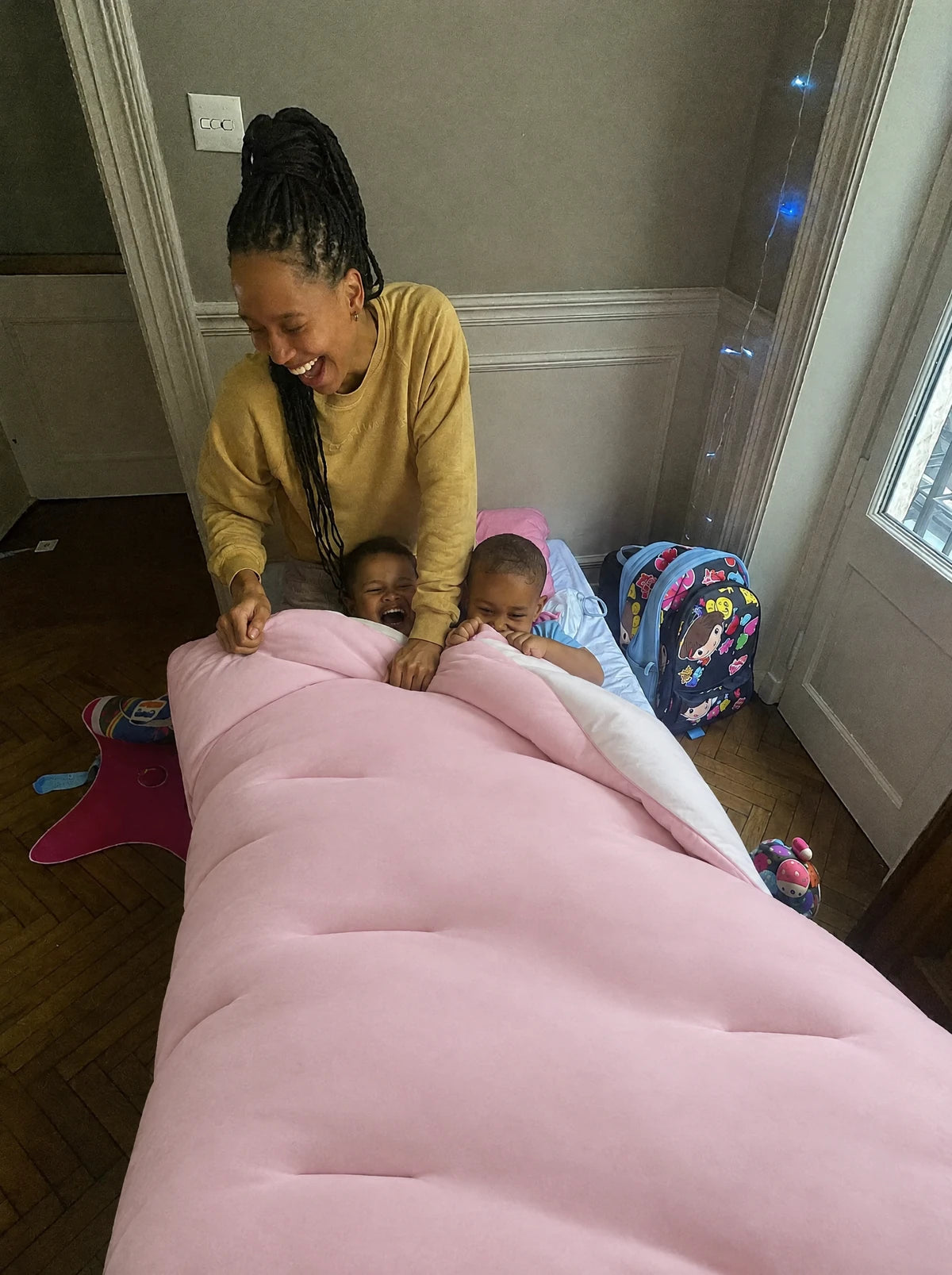 Deux enfants blottis sous une couette Whoosh rose poudré, leur maman assise au bord du lit